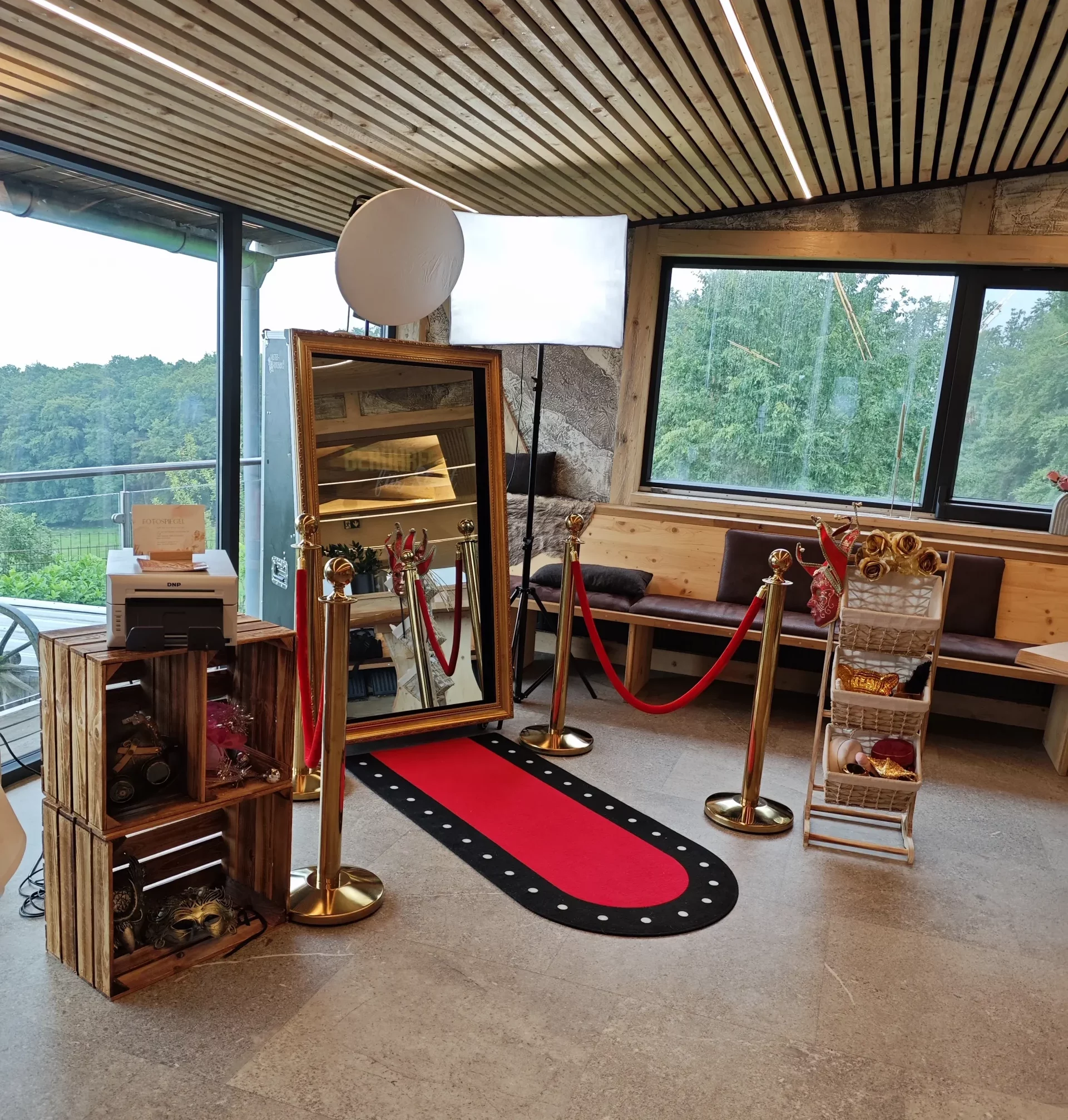 Fotospiegel mit goldfarbenem Rahmen, davor ein roter Teppich mit Absperrständern und roten Kordeln; daneben Requisitenregale und ein Drucker für Sofortausdrucke.